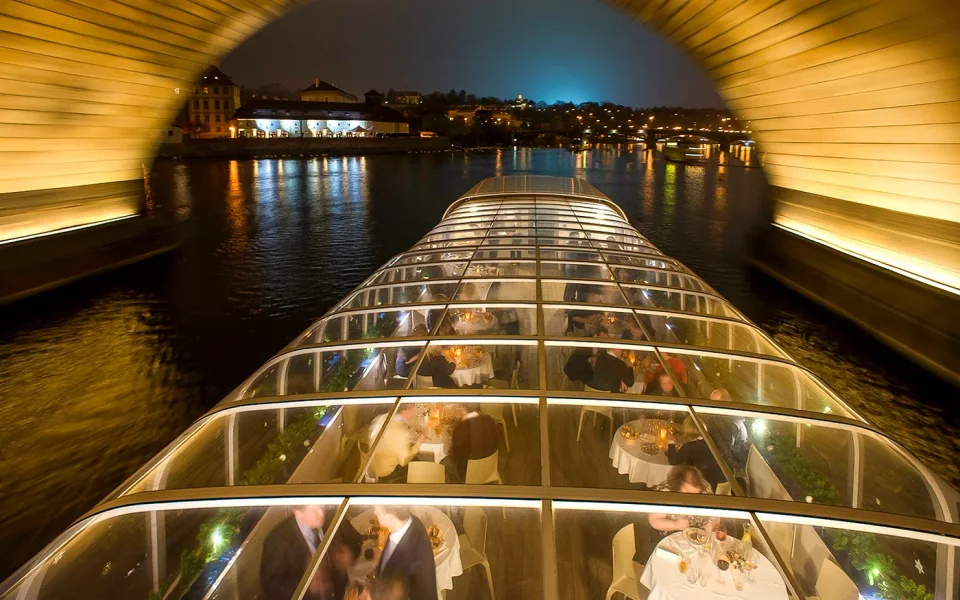 Illuminated glass-roofed dinner boat passing beneath a bridge